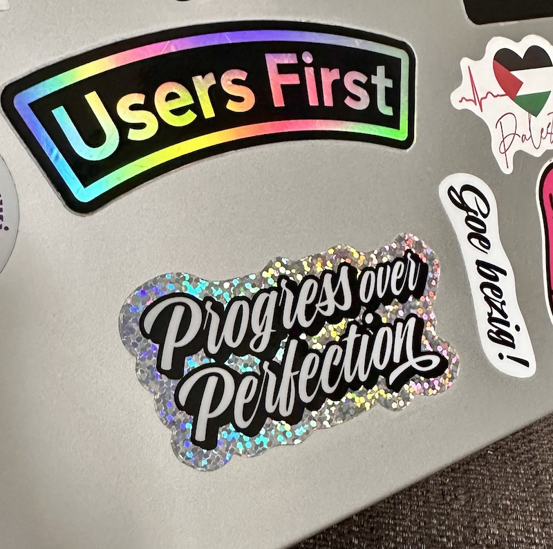 The “Progress over Perfection” sticker on a laptop, together with stickers that say “Users first” and “Goe bezig!”. There is also a sticker celebration the people from Palestine.