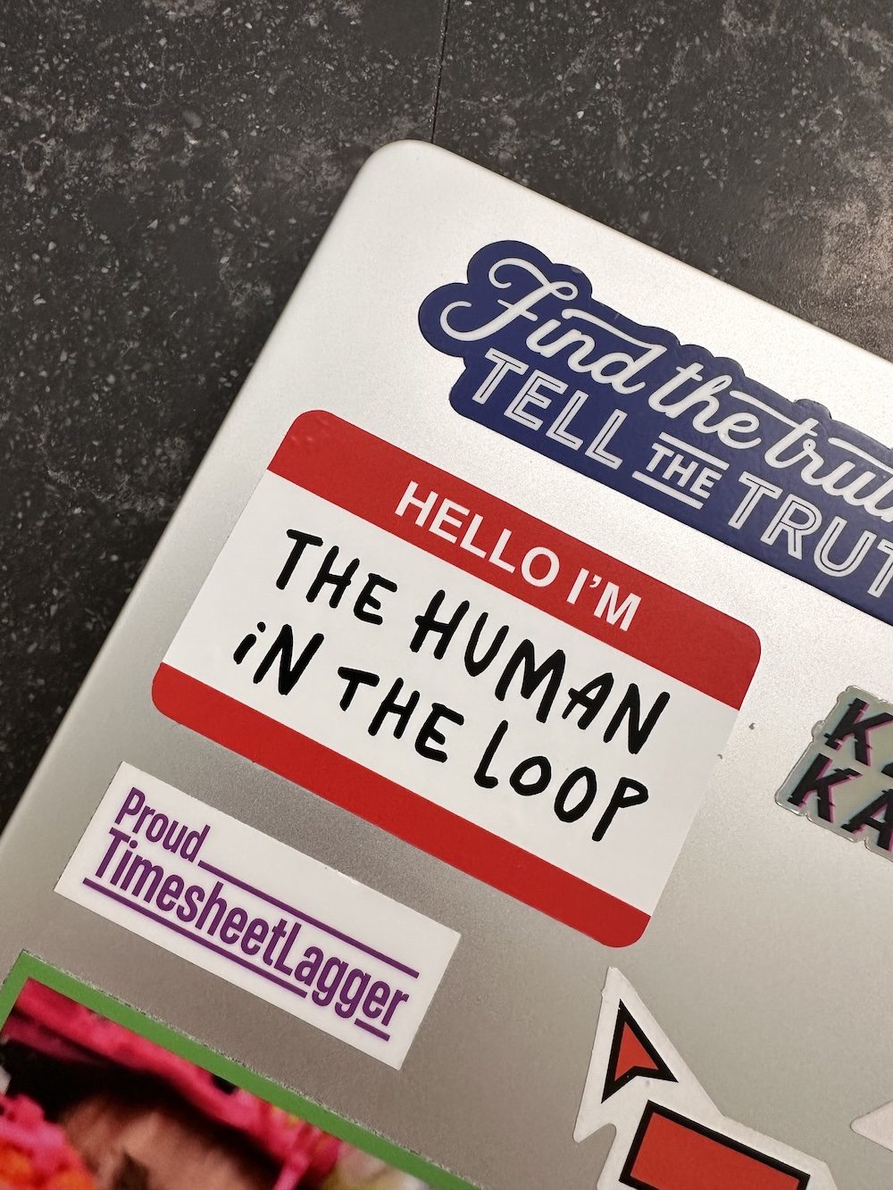 The ‘Human in the loop’ sticker on a laptop lid, with other stickers.